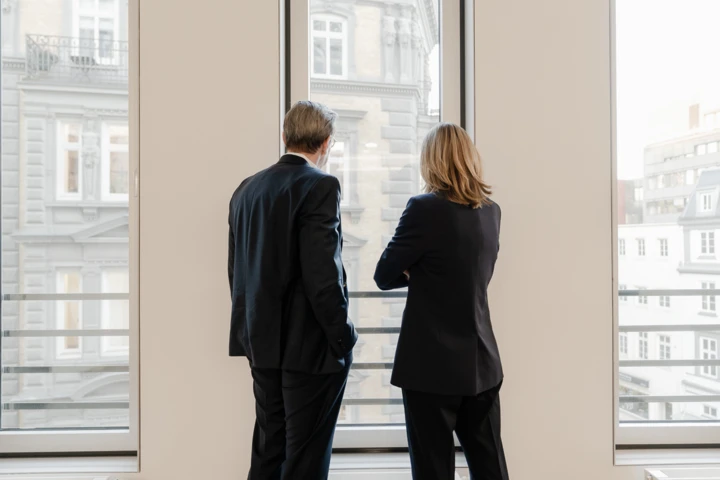 Zwei Personen stehen an bodentiefen Fenstern und schauen nach draußen.