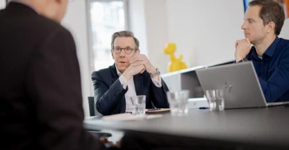 Drei Personen sitzen an einem Besprechungstisch mit Wassergläsern daruf.